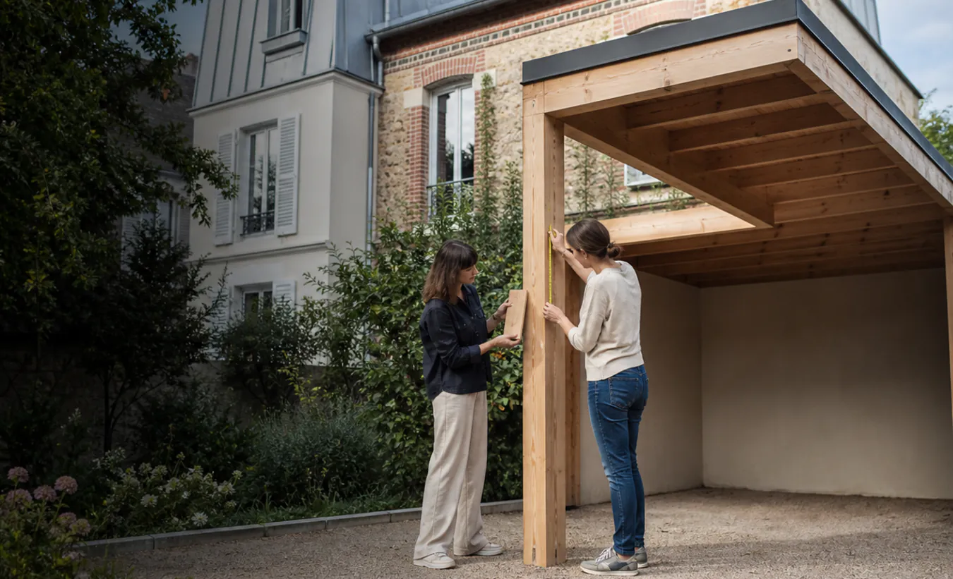 Une architecte et un propriétaire mesurent un carport en bois près d'une maison francilienne en brique et enduit clair