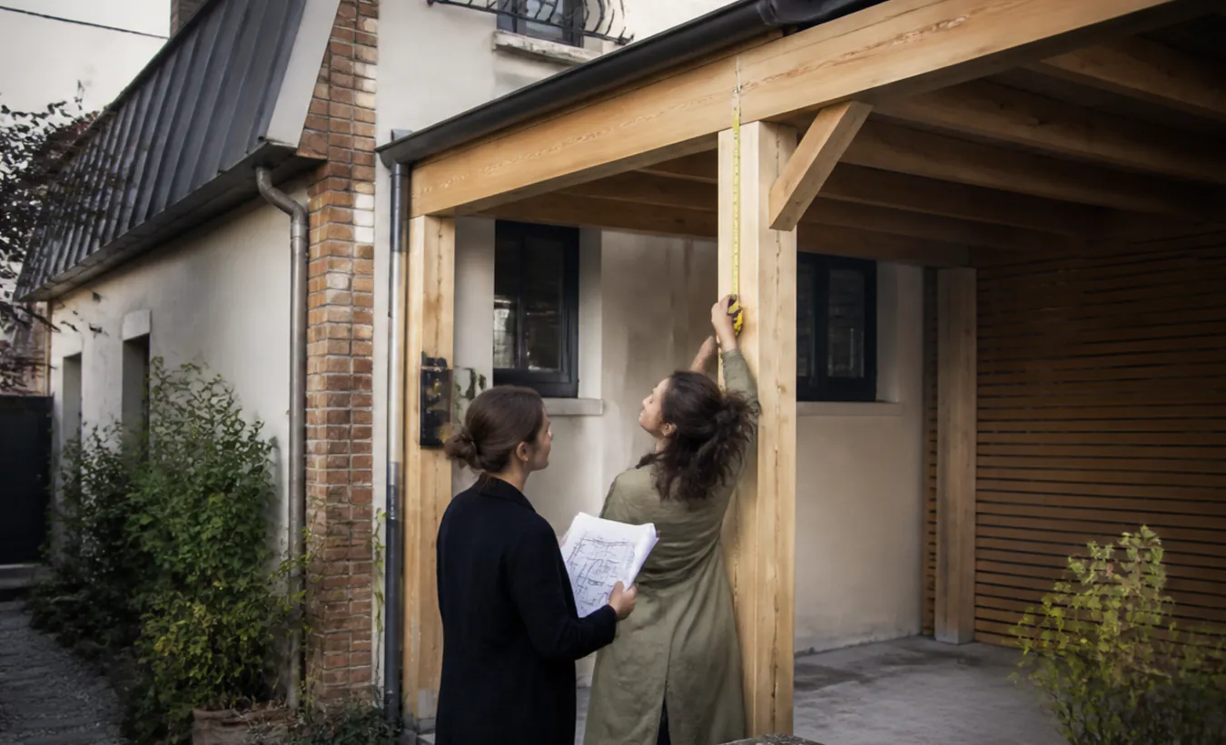 Une architecte et un propriétaire mesurent un carport en bois près d'une maison d'Île-de-France avec un jardin compact